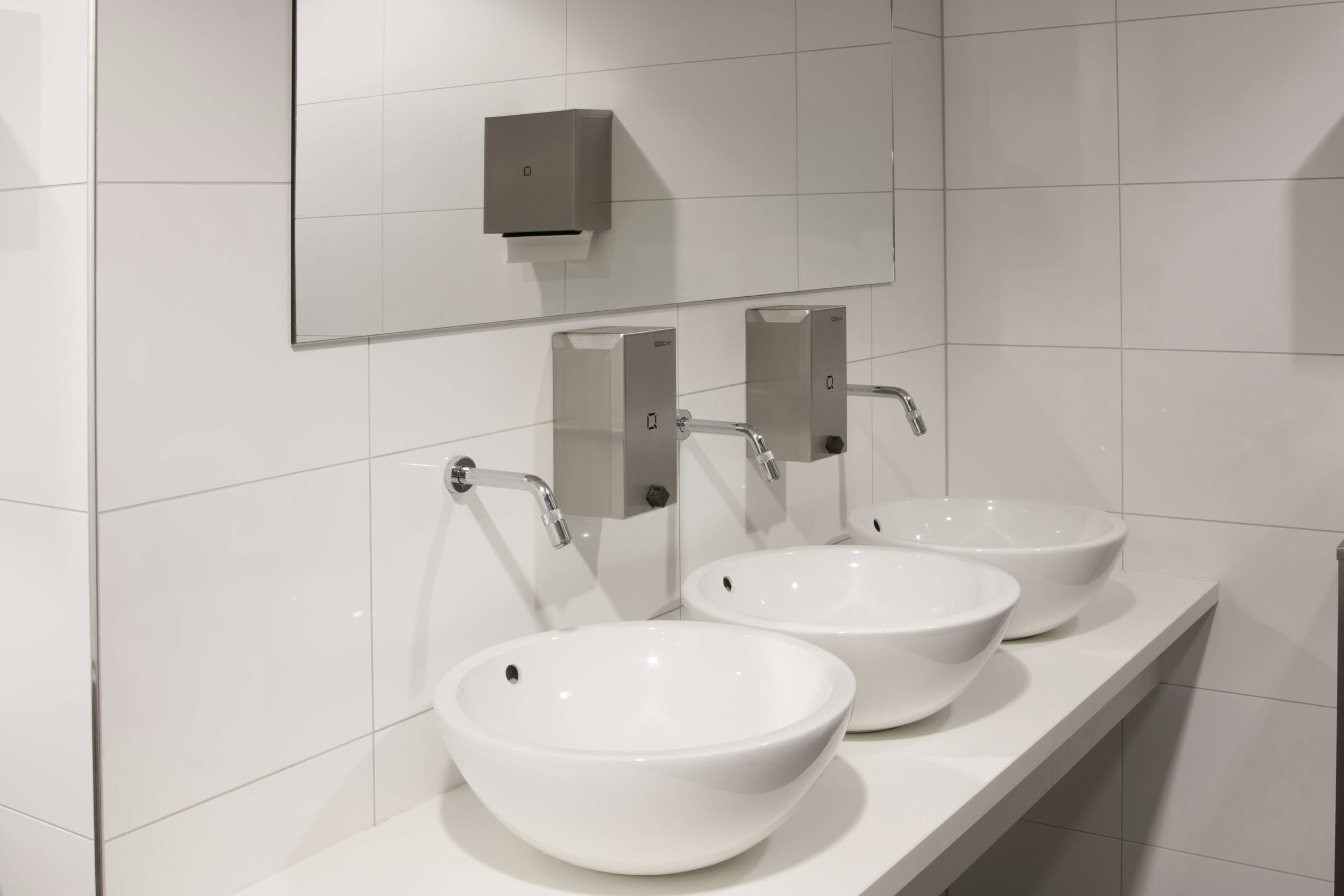 Drie witte wasbakken met zilveren kranen, twee zilveren zeepdispensers en een spiegel aan de muur.